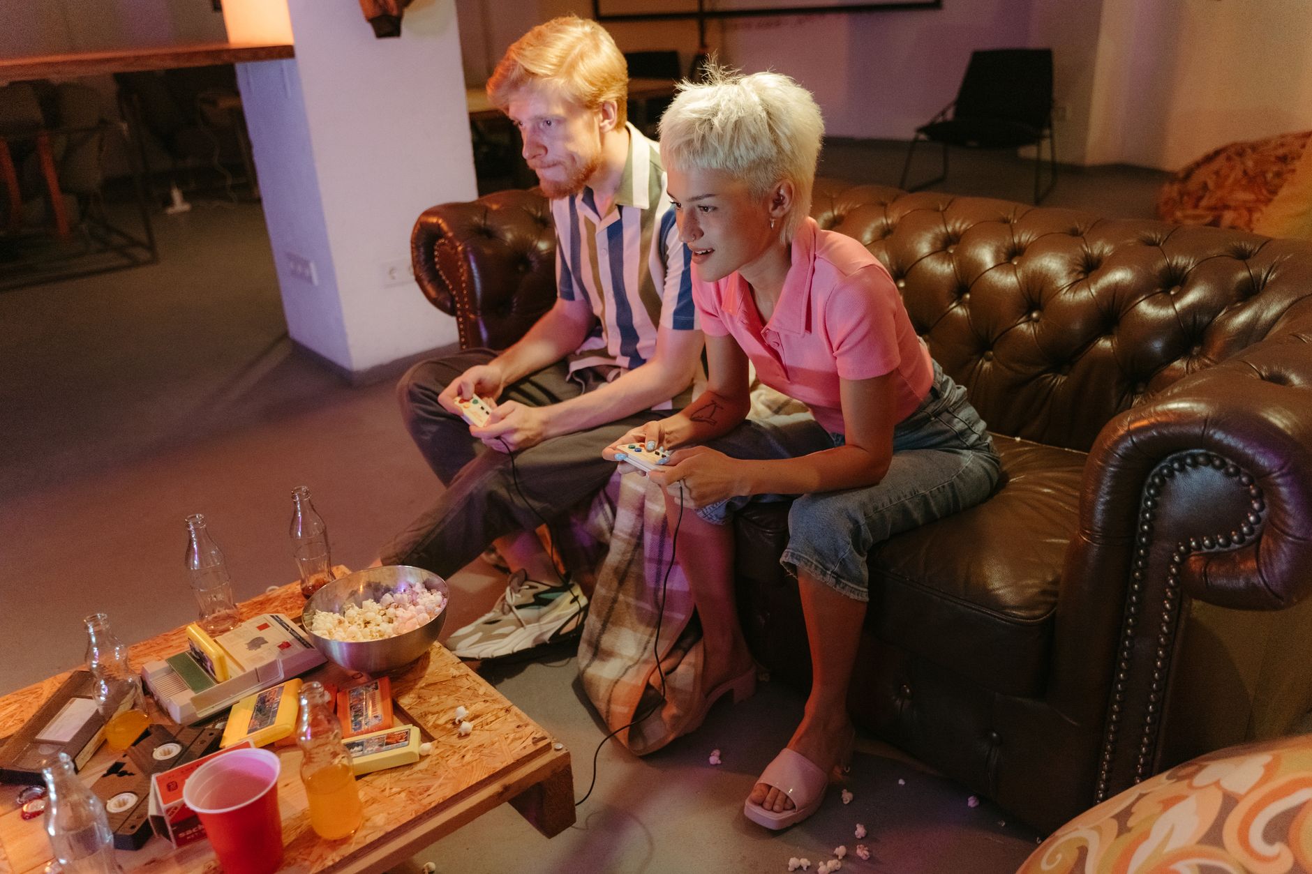 Deux amis jouant à des jeux vidéo sur un canapé en cuir, entourés de collations et de boissons, profitant d'une soirée de jeu décontractée.