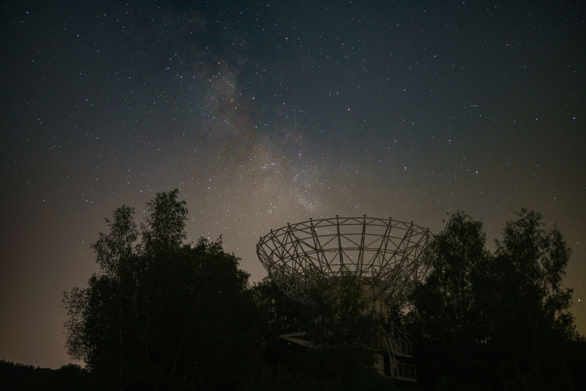 Antenne de radiotélescope en treillis entre des arbres, sous un ciel nocturne étoilé où se devine la Voie lactée.