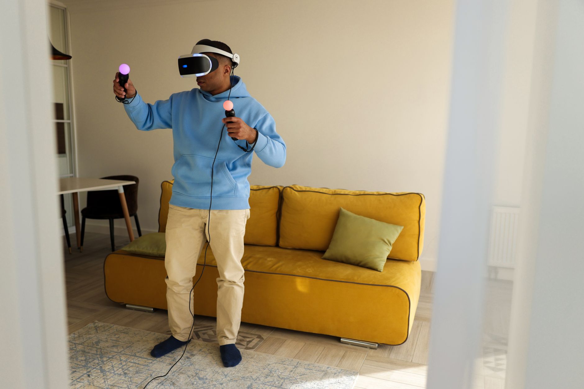 Homme en sweat bleu portant un casque de réalité virtuelle et tenant deux manettes lumineuses, debout dans un salon devant un canapé jaune.