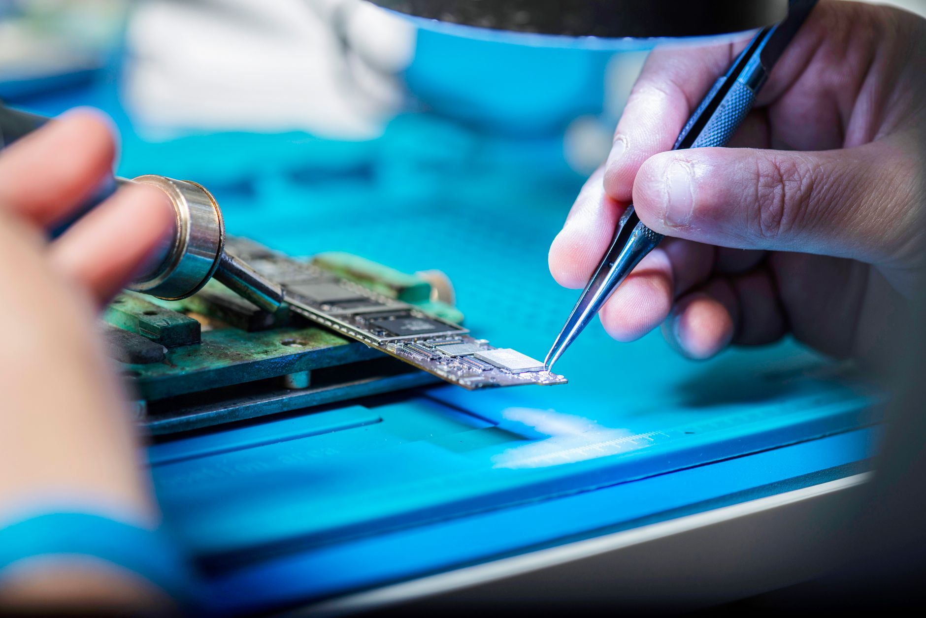 Manipulation d’une carte de circuit imprimé avec une pince fine et un fer à souder sur un plan de travail bleu.