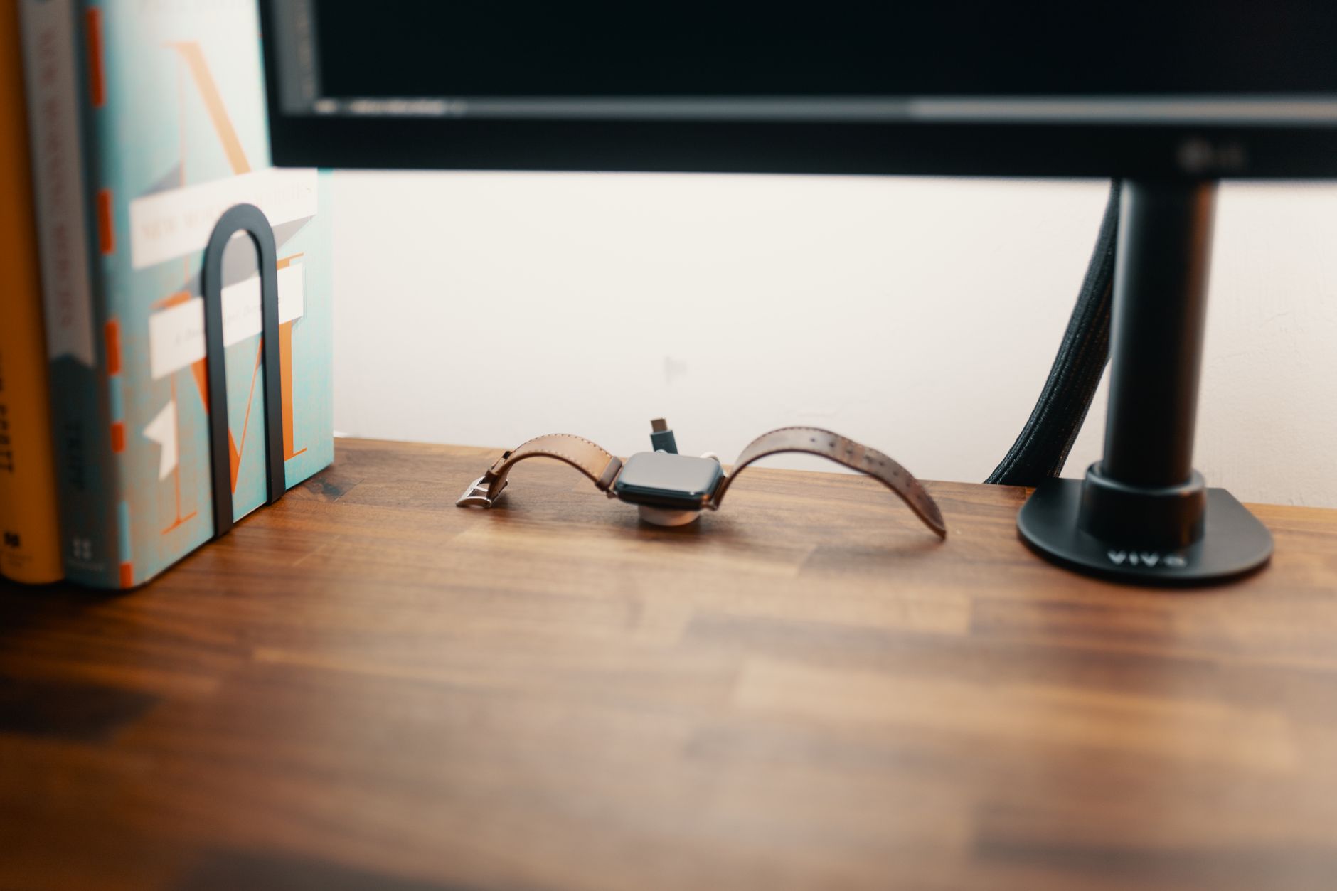 Montre connectée à bracelet en cuir marron en charge sur un bureau en bois, près de livres et du pied d’un écran.